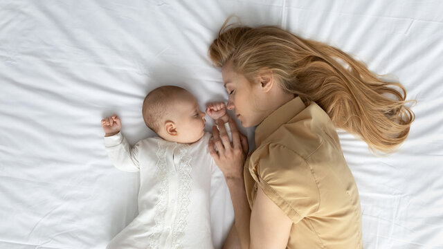 Tired mom and cute peaceful baby sleeping on mattress with white sheet. Calm young mother and sleepy infant resting in bed, enjoying relaxation, leisure time together. Child care concept. Top view.
