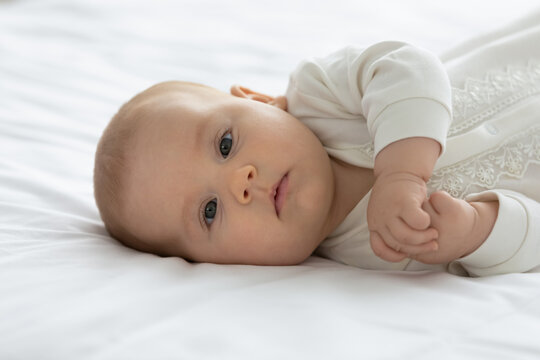 Little adorable few month baby lying on mattress, looking at camera. Cute infant boy or girl in bodysuit, resting in bed with white linen. Family, childhood, childbirth concept. Child portrait