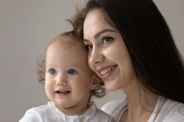 Close up portrait of happy new mom and cute blue eyed baby girl enjoying time together. Smiling young mother holding adorable toddler kid in arms, looking away. Motherhood, child care concept