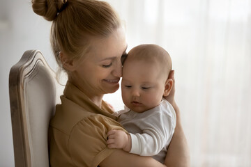 Happy peaceful mom hugging and kissing adorable baby, holding little kid in arms. New mother cuddling cute infant with love and tenderness, enjoying care of child, motherhood. Family, parent concept