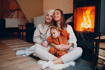 Grandmother, Mother and child sitting and playing on sofa near fireplace. Mom and baby. Parent with...