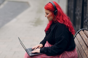 European plus size woman uses laptop while sitting on wooden bench outdoors at daytime. Young...