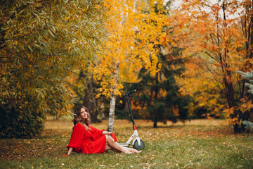 Young pretty woman with electric scooter in red dress sit on grass at the autumn city park.