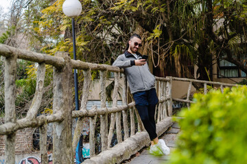 young hispanic latino man smiling leaning on a railing using a smart phone in a park outside, in autumn, wearing sportswear. copy space