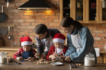 Smiling family couple with happy small kids wearing aprons, decorating gingerbread Christmas...