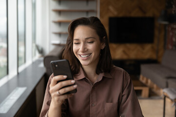 Fototapeta premium Smiling millennial Caucasian woman look at smartphone screen talk speak on video call online. Happy young female use cellphone have webcam digital virtual event on gadget. Communication concept.