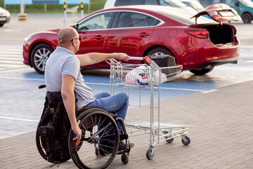 Fototapeta premium Adult disabled man in a wheelchair pushes a cart towards a car in a supermarket parking lot