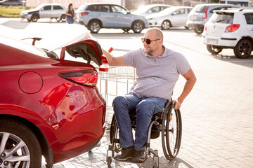 Adult disabled man in a wheelchair puts purchases in the trunk of a car in a supermarket parking lot
