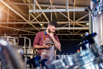 Fototapeta premium Young male brewer in leather apron supervising the process of beer fermentation at modern brewery factory