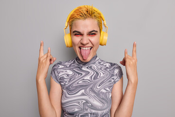 Sassy hipster girl with trendy yellow hair vivid makeup shows heavy metal horns gesture sticks out...