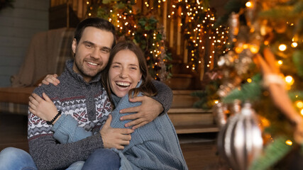 Fototapeta premium Portrait of happy emotional young affectionate family couple in warm winter sweaters cuddling in decorated living room, feeling excited of celebrating Christmas or New year together, holidays mood.
