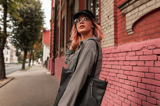Hipster beautiful young woman with glasses and a cap in fashionable youth clothes with a bag walks in the city near a vintage brick building