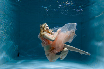 A girl with long dark hair swims underwater in a pink dress and with a crown on her head, like an...
