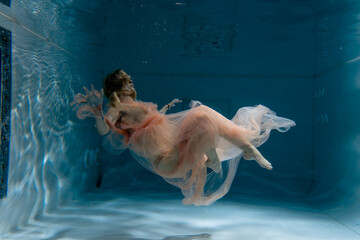 A girl with long dark hair swims underwater in a pink dress and with a crown on her head, like an underwater queen. Fairy tale suitable for advertising	
