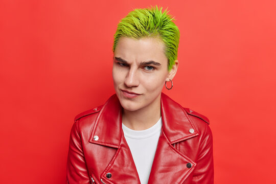 Photo of hipster girl with trendy green hairstyle belongs to youth subculture has unique style. Trendy serious female rapper in youth style clothing isolated over vivid red background. Adolescence