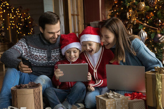 Happy Young Parents Sitting On Floor Near Decorated Christmas Tree, Watching Funny Comedian Movie Or Cartoons With Adorable Small Children On Digital Tablet, Unpacking Gifts Together In Living Room.