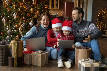 Technology addicted overjoyed young family couple watching funny cartoons online or playing games on touchpad with happy sincere small adorable children siblings, unwrapping Christmas gifts at home.
