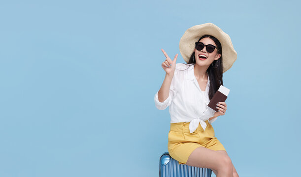 Happy smiling Asian woman dressed in summer clothes and wearing hat with luggage enjoying their summer vacation and pointing finger a copy space on bright blue background..