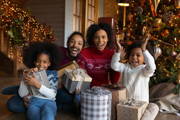Portrait of overjoyed young African American family with two small children have fun open presents on Christmas morning at home. Happy parents with little kids celebrate New Year winter holidays.