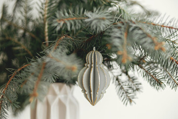 Merry Christmas! Stylish christmas ornament on fir branch in modern vase close up. Modern wooden bauble on branch on white background. Festive simple decor in scandinavian room.
