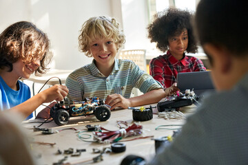 Happy cute schoolboy looks at camera sits at desk with group of diverse middle school students building innovative robot making software engineering at science lab class. STEM technologies concept. - Powered by Adobe