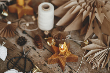 Stylish Christmas candle, golden bells, white ornaments, pine cones, paper stars, thread and scissors on rustic wooden background. Making festive scandinavian garland, moody image