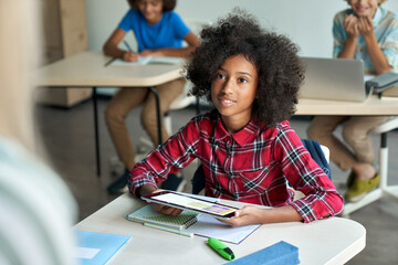 Happy African American schoolgirl looking listening to teacher using tablet device sitting in classroom with group of schoolchildren using laptop computers. Modern technologies for education concept.