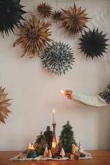 Hand lighting up christmas candle on background of pine trees and cones on wooden table and paper...