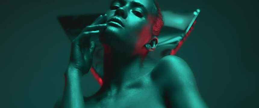 Portrait of young woman with shiny makeup and skin in colorful neon light. Female model moving sensually looking at camera in studio. Futuristic advertisement, modern fashion, concept. Anamorphic shot