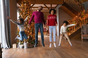 Portrait of smiling young African American family with kids jump dance near fir tree on Christmas...