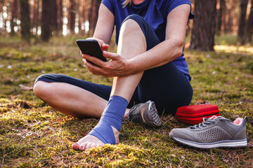 Injured hiker with sprain ankle using smart phone to get help. Woman gave herself first aid during hiking injury in nature - obrazy, fototapety, plakaty