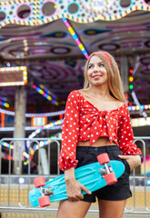 Cute cheerful blonde woman with penny board having fun in the amusement park