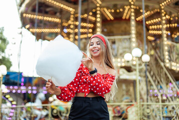 Obraz na płótnie Canvas Adorable cute beautiful woman with cotton candy stands in the middle of an amusement park with bright colors, positive and cheerful, happy and optimistic emotion