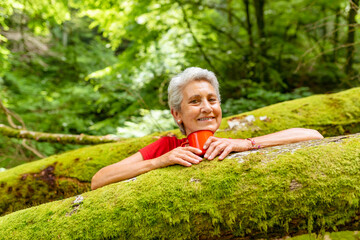 Happy 70 year old woman holds a red cup with drink while smiling looking at camera on a beech log...