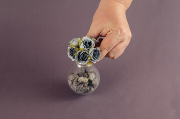 A woman's hand holds an electric lamp with artificial flowers on a gray background. Gray roses inside a glass vessel with gravel. Selective focus.