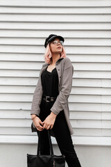 Fashion stylish beautiful young woman with glasses and a hat in trendy casual clothes with a fashion bag stands near a wooden wall