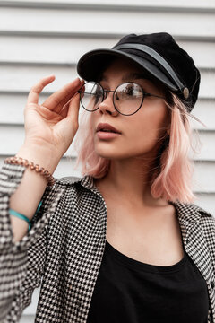 Cool fashion portrait of a hipster pretty young teenager woman with casual fashionable outfit puts on and straightens vintage round glasses near a wooden wall