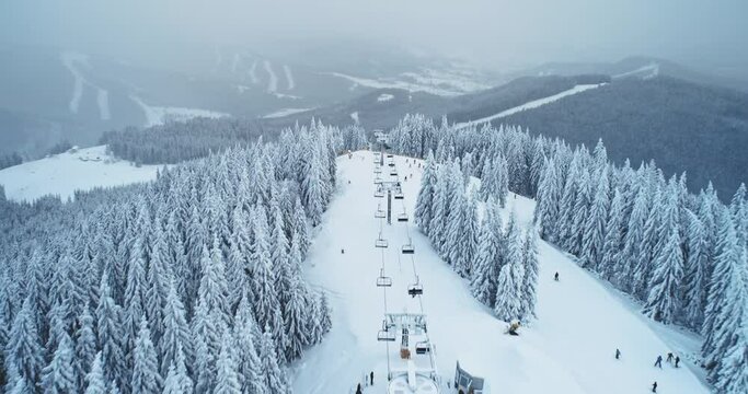 Aerial winter ski lift in mountain forest, skier going down. Flight over pine trees in white snow, slow motion. Relax for downhill skiing. Snow slopes at ski resort. Outdoor tourism. Cinematic shot