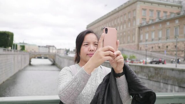 Asian woman standing and taking a picture of river and town in Sweden, traveling abroad on holiday.Walking on the bridge and using smartphone to take a photo. Beautiful town in Sweden