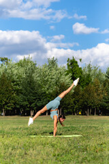 A beautiful sportive girl in sportswear practices yoga on the green grass in the city park. Handstand with twine