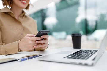 Smiling young entrepreneur woman sitting in cafe outside with phone in hands in front of open laptop