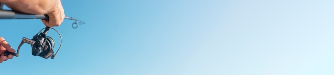 Fishing rod reel in fisherman hands over clear blue sky with copy space for text. Long wide horizontal web banner with copyspace.
