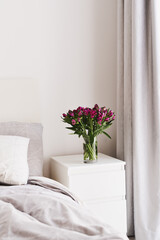 Simple home decor: fresh Alstroemeria flowers in a glass vase on a bedside table of white color in a bedroom. Cozy and comfortable atmosphere. Minimal style in interior design