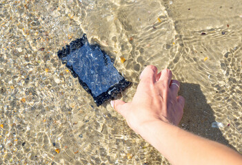 A woman's hand lifts a her smartphone from sea water on the beach. Lost and found accessories or...
