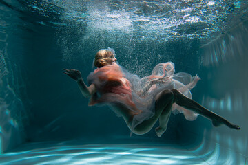A girl with long dark hair swims underwater in a pink dress and with a crown on her head, like an...