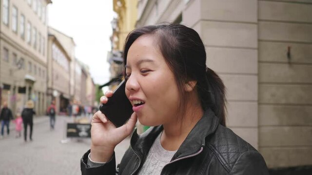 Happy Asian woman standing and having a call on street in Sweden, going out for a walk on the street in small town. Traveling abroad on long holiday.