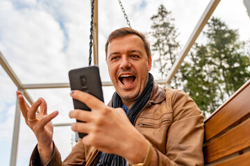 Man with happy face checks his phone while sitting on wooden swinging bench in autumn park...