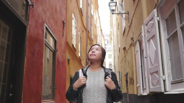 Front view of happy Asian woman walking on street in Sweden, going out for a walk on the street in small town. Traveling abroad on long holiday. Colorful building background