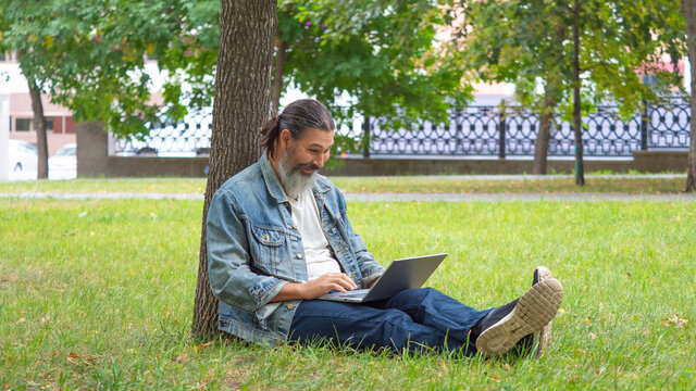 Middle aged man with laptop in city park. He sitting on the grass in the park under the tree. Format photo 16x9.