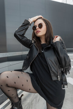 Beautiful sexy model woman with stylish round sunglasses in a fashionable leather jacket and black sweatshirt with sexy tights holds a bag and posing on the street